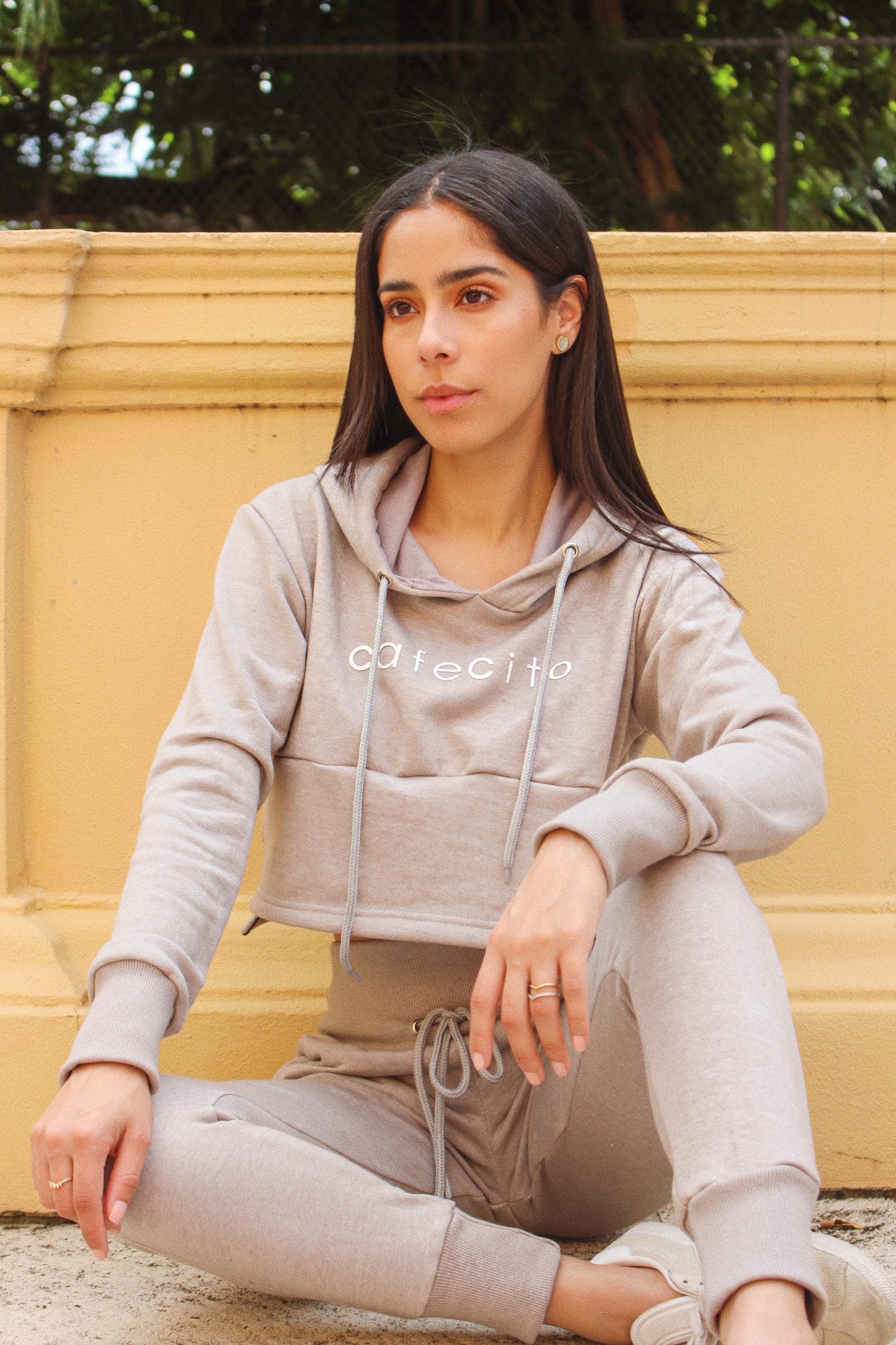 Woman Sitting in Grey French Terry Drawstring Hoodie and Pant Set with cafecito screen next to a yellow wall and tree